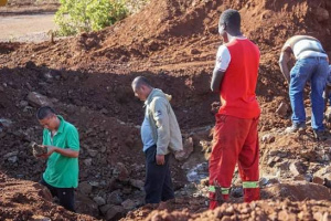 Four men stand and dig in a large pit of reddish soil at what appears to be a small-scale mining site, examining rocks and working with tools as they search for minerals.